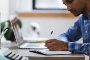 a man blogger writing a blog post fast in a book with a laptop on the table